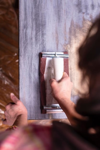Close-up of a person preparing a wooden window frame for painting. They are using sandpaper to smooth the surface and remove old paint flakes. The background shows the rest of the house with a partly faded paint job.