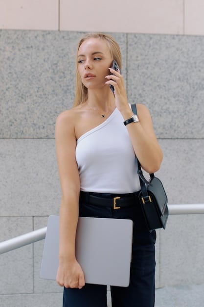 A professional woman in a grey blazer and white blouse, adorned with a sleek metallic watch, delicate stud earrings, and a structured tote bag, sitting at a modern desk with a laptop.
