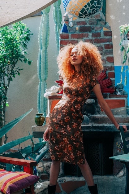 A woman with long curly hair wearing a layered bohemian outfit, including a printed maxi skirt, a graphic tee, a denim jacket, and several long necklaces, smiling brightly and posing in an urban park.